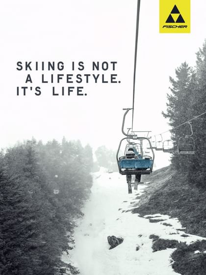 A skier sits alone on a chairlift in falling snow, surrounded by snowy trees – featuring the slogan “Skiing is not a lifestyle. It’s life.” and the Fischer logo.