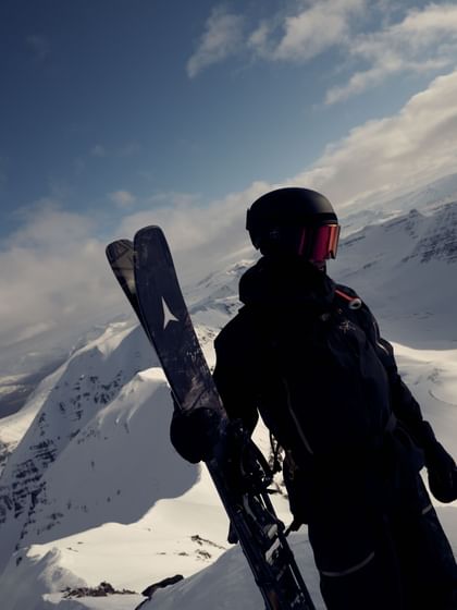 Ein Freerider in schwarzer Skibekleidung, Helm und rot verspiegelter Skibrille hält seine Atomic Ski aufrecht neben sich. Im Hintergrund erstreckt sich eine verschneite Berglandschaft unter blauem Himmel.