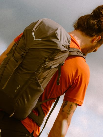 Ein Mann mit Dutt und rotem Shirt trägt einen schwarzen Wanderrucksack auf einem Bergpfad. Die Szene ist aus der Rückenperspektive aufgenommen und vermittelt Fokus und Bewegung.