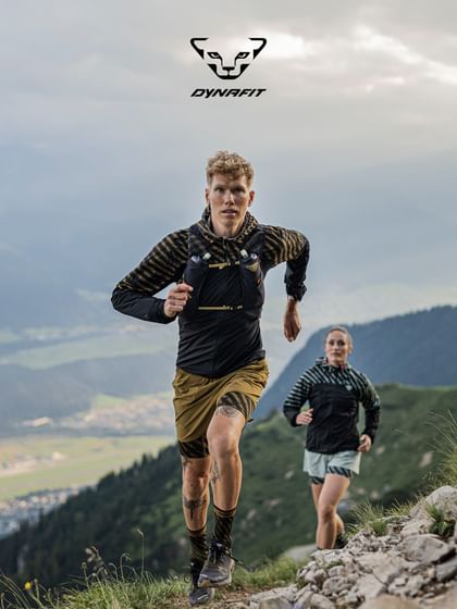 Zwei Trailrunner laufen auf einem steinigen Bergpfad mit atemberaubendem Panorama im Hintergrund. Der Mann im Vordergrund trägt Dynafit-Laufkleidung in Schwarz-Gold, die Frau folgt dicht dahinter. Beide sind konzentriert und energiegeladen in alpiner Umgebung unterwegs. Oben rechts ist das Dynafit-Logo zu sehen.