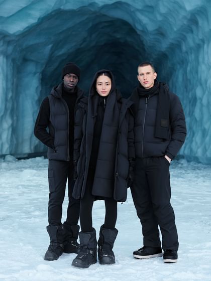 Drei Personen stehen in AlphaTauri Winterkleidung vor einer blauen Eisgrotte, alle tragen schwarze Jacken, Hosen und Winterstiefel.