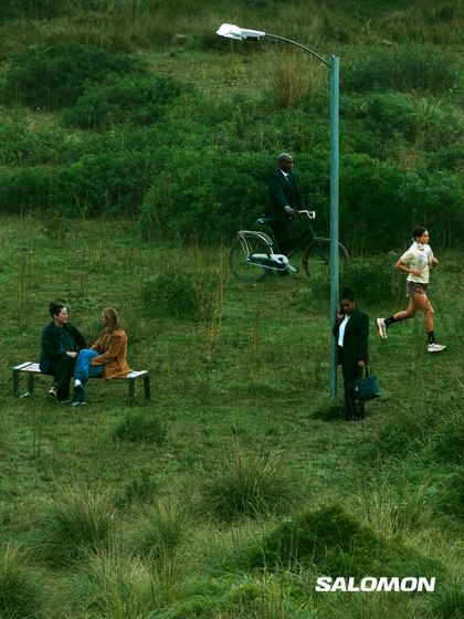 Several people move through a grassy natural landscape, including a runner, a cyclist, and people in business attire; the Salomon logo appears in the bottom right corner.