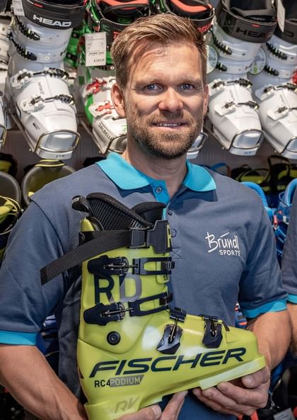 Das Bild zeigt einen Mitarbeiter von Bründl Sports, der ein Paar gelbe Fischer RC4 Podium Skischuhe in den Händen hält. Im Hintergrund sind verschiedene Skischuhe von Marken wie Head und Dalbello auf Regalen zu sehen. Der Mitarbeiter trägt ein dunkelblaues Polohemd mit dem Bründl Sports Logo.