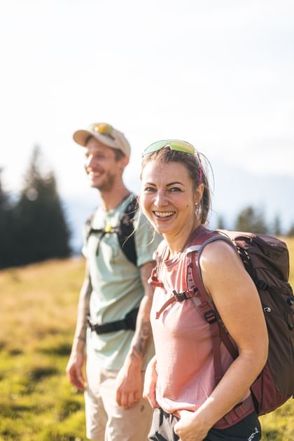 A woman wearing a backpack and sunglasses on her head smiles at the camera, while a man with a cap and backpack stands slightly blurred in the background. They are on a sunny meadow with trees and mountains in the background.