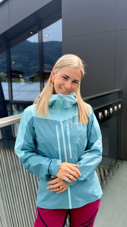 A woman with blonde hair stands on a balcony in front of a modern building with a glass facade, smiling at the camera. She is wearing a light blue Norrona outdoor jacket and pink pants, with mountains and houses visible in the background.
