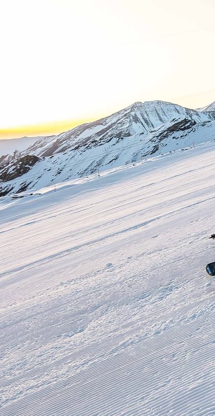 A skier carves through a freshly groomed slope surrounded by snow-covered mountains with the rising sun in the background.