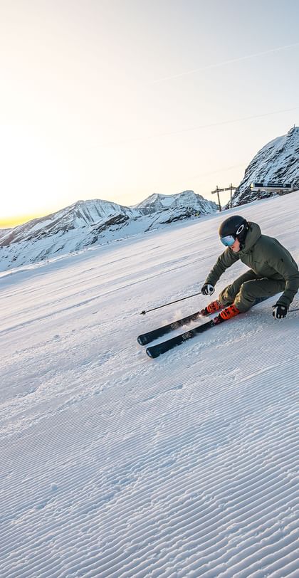 Das Bild zeigt einen Skifahrer, der bei Sonnenaufgang eine schneebedeckte Piste hinunterfährt, umgeben von Berglandschaft.