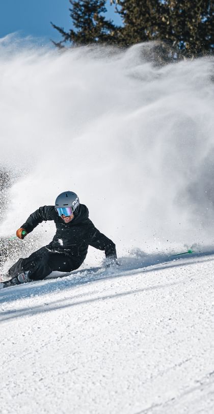 Ein Skifahrer in schwarzer Skibekleidung fährt in tiefer Schräglage über die Skipiste und wirbelt eine große Schneewolke auf. Im Hintergrund sind Nadelbäume und blauer Himmel zu sehen.