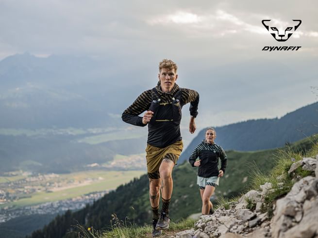 Zwei Trailrunner laufen auf einem steinigen Bergpfad mit atemberaubendem Panorama im Hintergrund. Der Mann im Vordergrund trägt Dynafit-Laufkleidung in Schwarz-Gold, die Frau folgt dicht dahinter. Beide sind konzentriert und energiegeladen in alpiner Umgebung unterwegs. Oben rechts ist das Dynafit-Logo zu sehen.