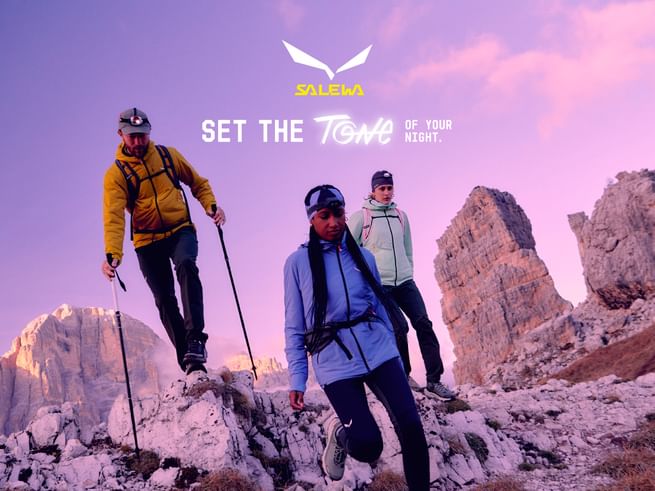 Three hikers with headlamps and trekking poles explore rocky alpine terrain at dusk. They are wearing colorful outdoor gear by Salewa. In the background, the mountains and sky glow in warm purple and pink hues.