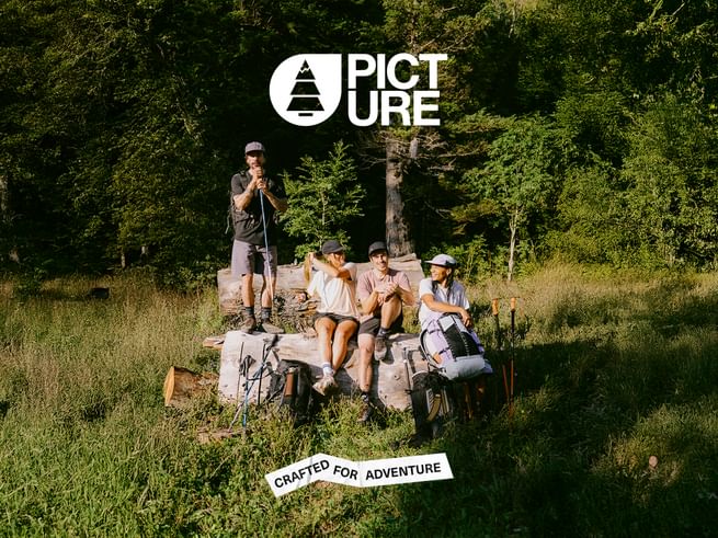 A group consisting of 2 women and 2 men in Picture Organic Clothing outdoor clothing take a break on a large tree trunk in a forest clearing. 