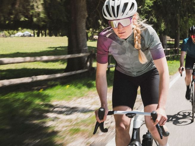 A focused cyclist in athletic clothing rides on a sunny country road, followed by another cyclist.
