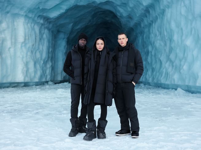 Drei Personen stehen in AlphaTauri Winterkleidung vor einer blauen Eisgrotte, alle tragen schwarze Jacken, Hosen und Winterstiefel.