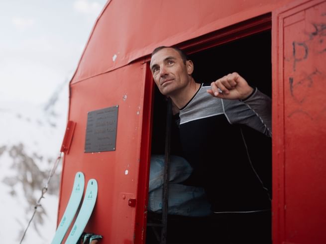 Real moments in the mountains – with Löffler, functional and mindful. A man looks out from a red mountain hut into the snowy alpine landscape. Light blue skis stand next to the door, featuring Löffler apparel.