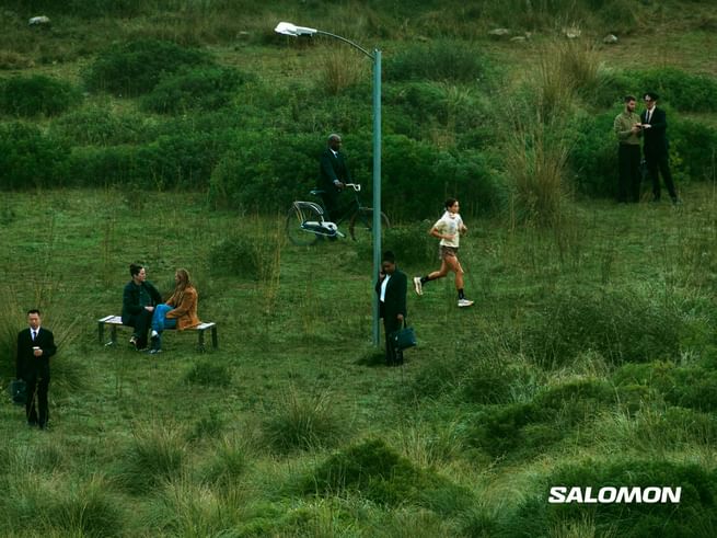 Several people move through a grassy natural landscape, including a runner, a cyclist, and people in business attire; the Salomon logo appears in the bottom right corner.