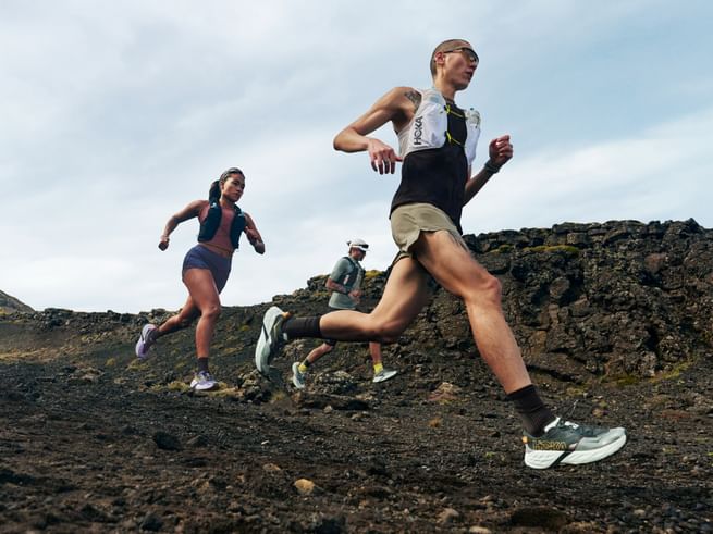 Drei Trailrunner laufen über einen felsigen Bergpfad und tragen Hoka Trailrunning-Schuhe sowie Laufwesten in rauer alpiner Landschaft.