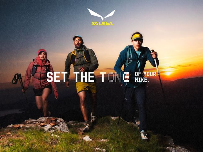 Three people wearing backpacks and outdoor clothing walk along a grassy, rocky mountain path as the sun sets on the horizon. The image includes the text “SET THE TONE OF YOUR HIKE.” and the logo “SALEWA”.