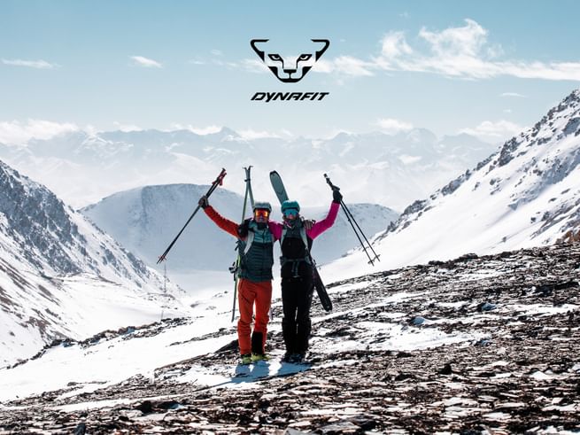 Two ski tourers celebrate their ascent in the snowy Alps, holding touring skis and ski poles, with the DYNAFIT logo visible above them.