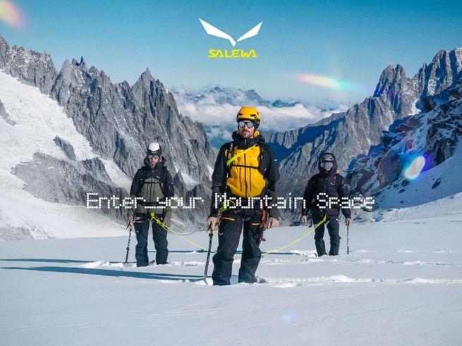 Three mountaineers connected by a rope, wearing helmets and backpacks and carrying poles, stand in snow beneath steep rocky peaks. The image features the Salewa logo and the text “Enter your Mountain Space”.