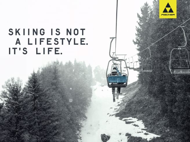 A skier sits alone on a chairlift in falling snow, surrounded by snowy trees – featuring the slogan “Skiing is not a lifestyle. It’s life.” and the Fischer logo.